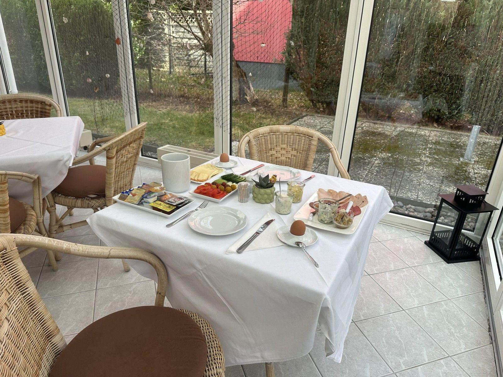 Glasfront mit Blick auf Garten. Tisch mit Frühstück und Stühlen.