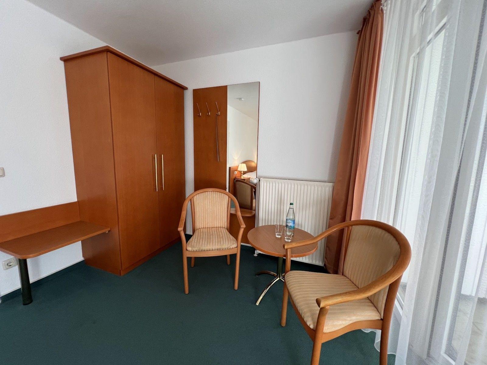 Zimmer mit Schreibtisch, Kleiderschrank, zwei Stühlen und Tisch. Fenster mit Vorhängen.