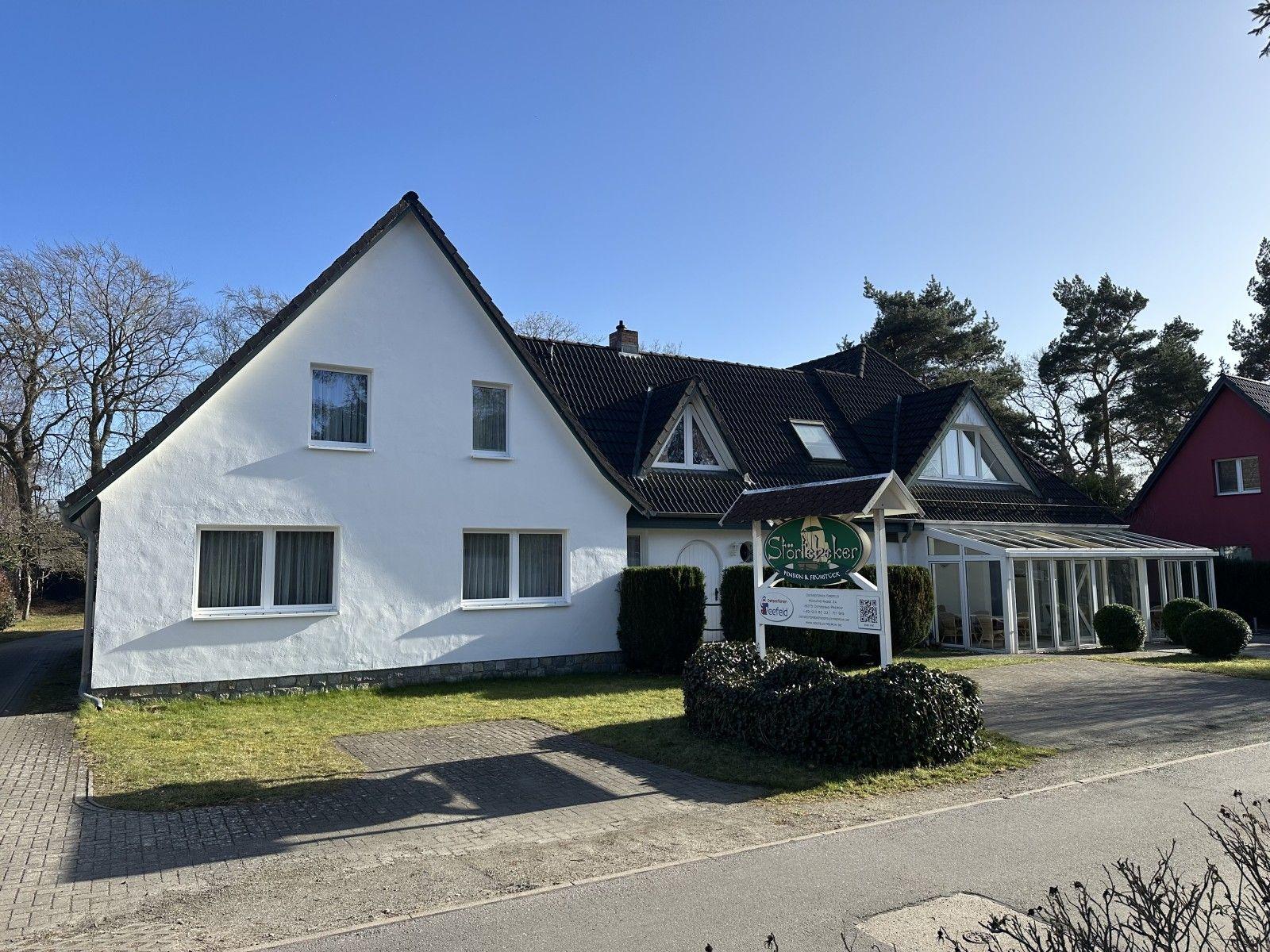 Weißes Haus mit dunklem Dach, Vorgarten und Schild. Neben einem roten Haus.