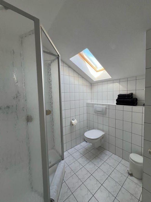 Badezimmer mit Dusche, Toilette und Dachfenster. Weiße Fliesen und Handtücher.