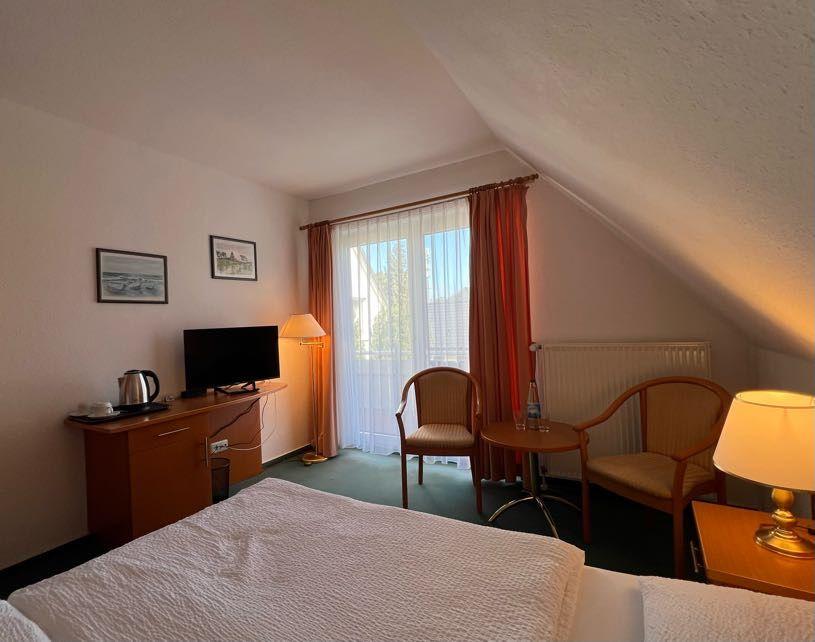 Zimmer mit Bett, TV, Schreibtisch und Sitzgelegenheit neben Fenster mit Vorhängen.