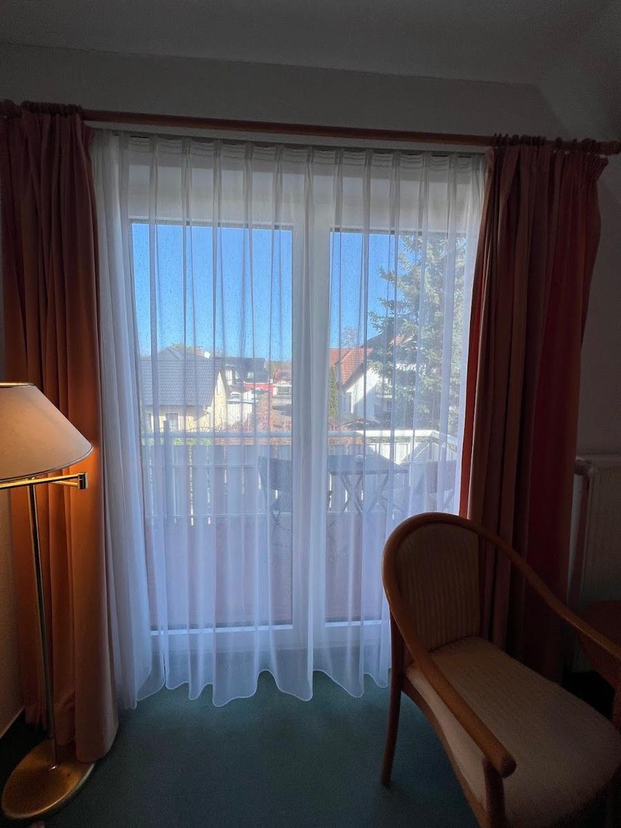 Zimmer mit Fenster, weißen Vorhängen und roten Gardinen. Draußen ist ein Balkon mit Tisch und Stühlen.