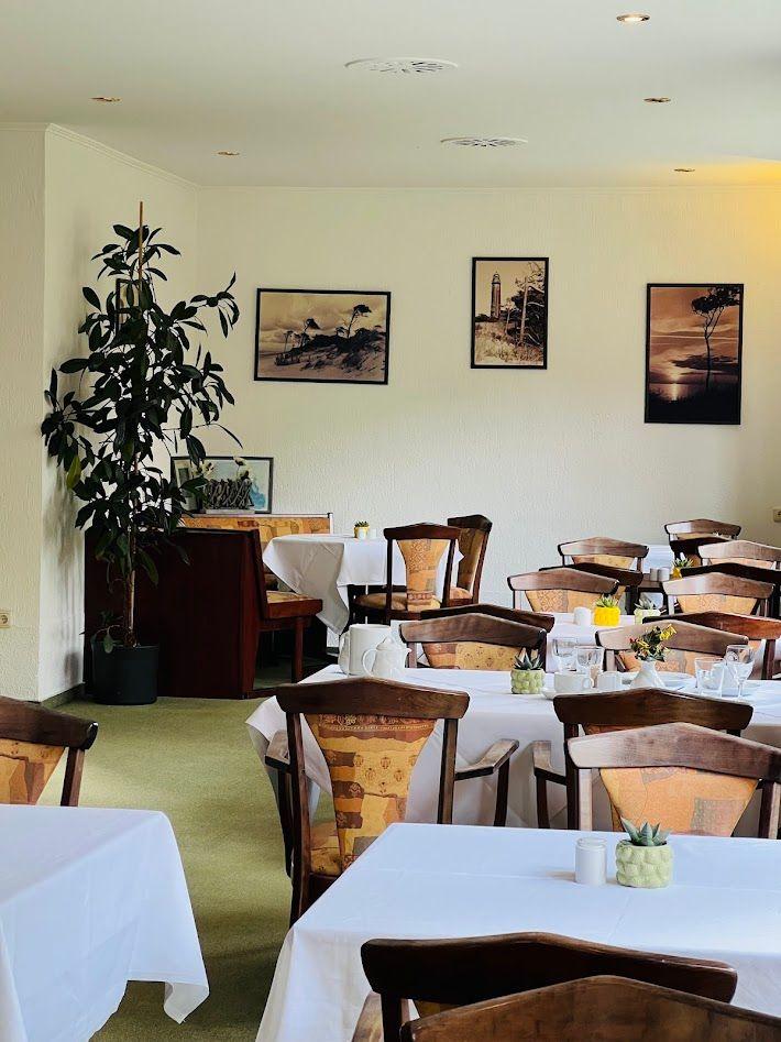 Esszimmer mit Tischen, Stühlen und Wandbildern. Grüner Teppich und Pflanze.