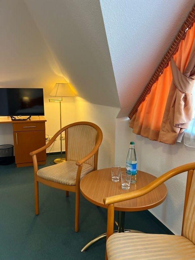 Zimmer mit Holzstühlen, runder Tisch, Fernseher und Fenster mit orangefarbenen Vorhängen.