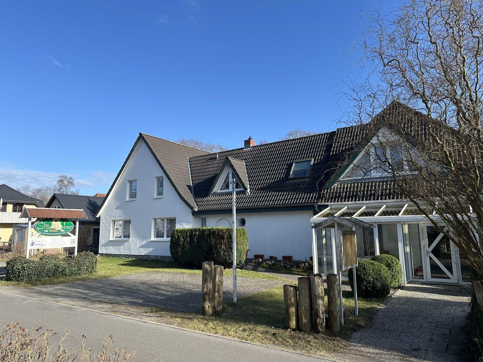 Weiße Ferienhaus mit Dachfenstern und Glasveranda. Grün bewachsenes Grundstück mit Holzpfosten.