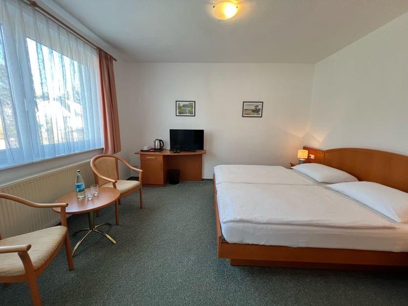 Zimmer mit Doppelbett, Schreibtisch, Fernseher und Sitzgelegenheit neben Fenster.