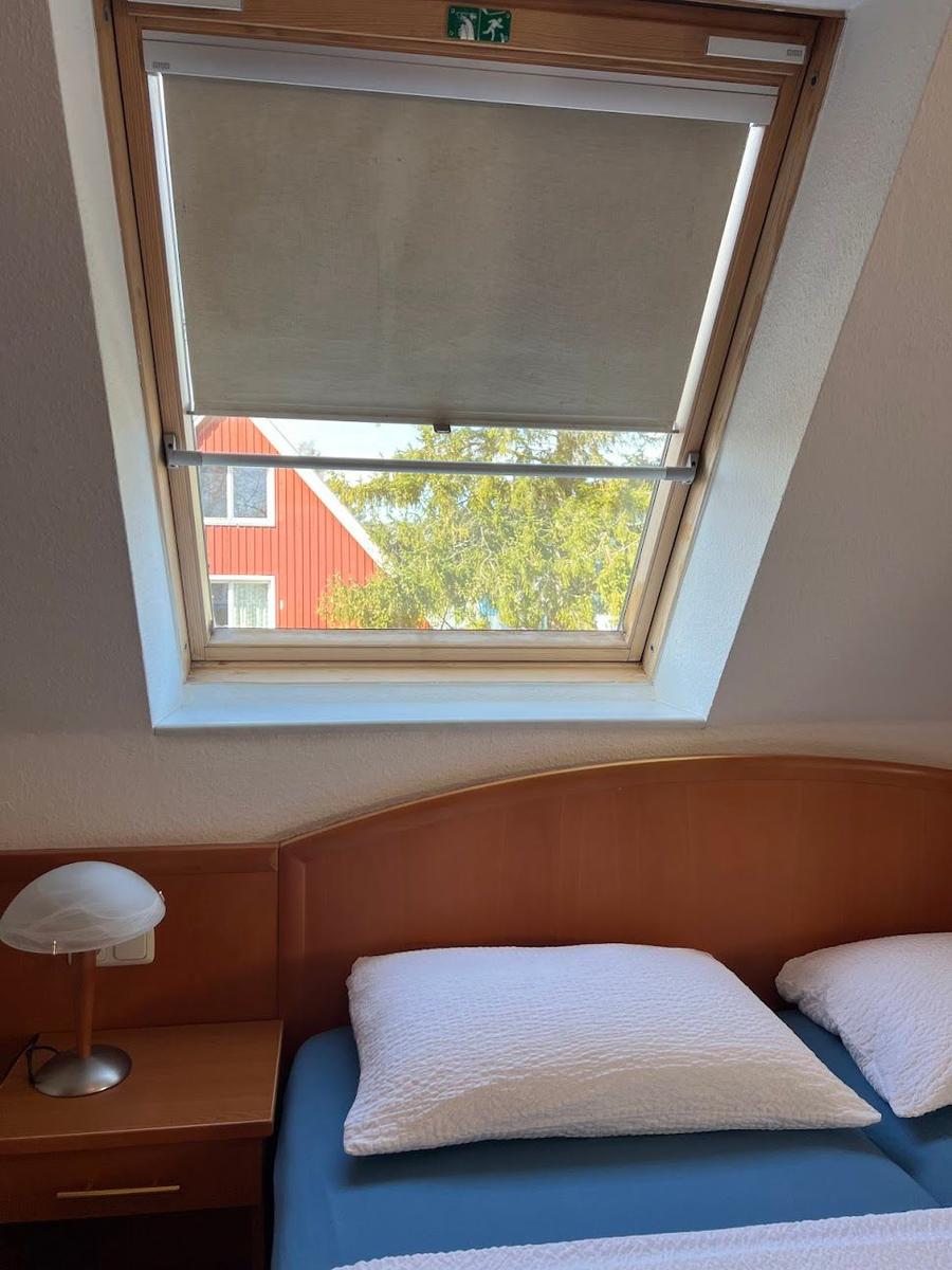 Schlafzimmer mit Bett, Nachttisch und Fenster. Draußen ist ein rotes Haus und Bäume sichtbar.