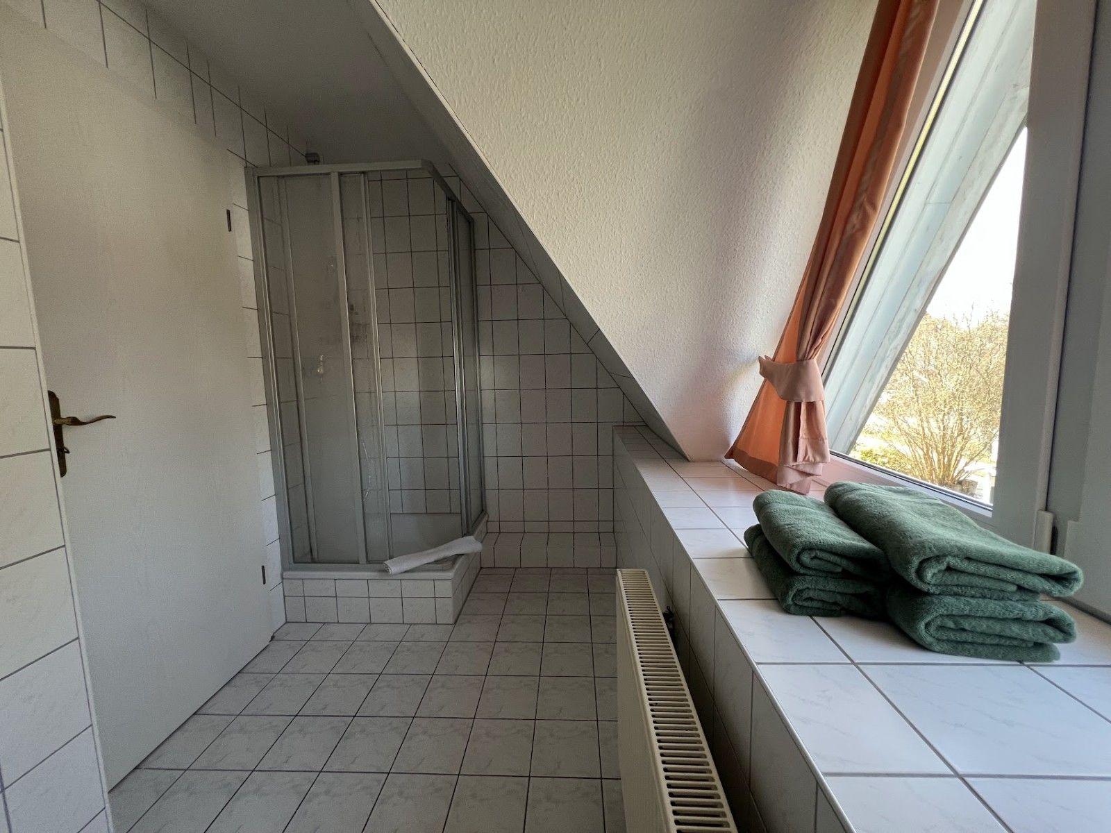 Badezimmer mit Dusche, Fenster und grünen Handtüchern auf Fensterbank.