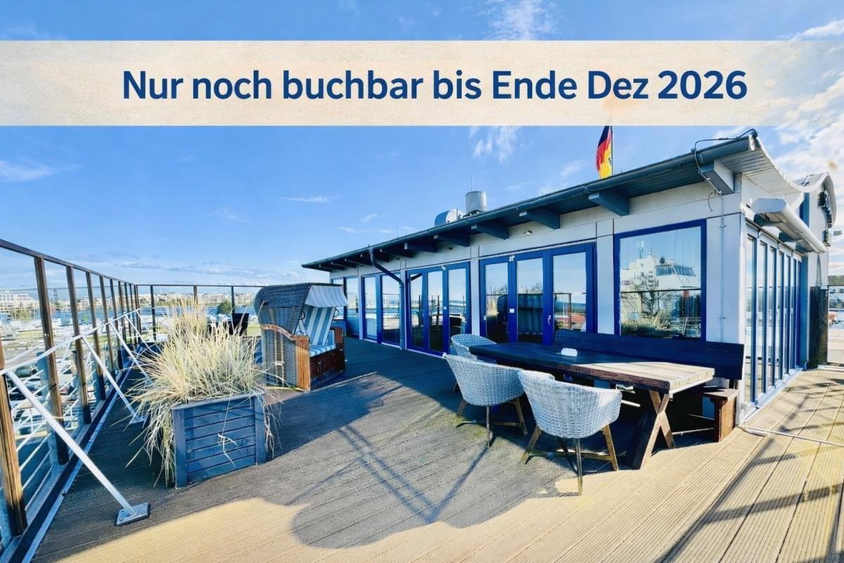 Terrasse mit Tisch, Stühlen und Liegehaus am Wasser. Gebäude mit blauen Fenstern.