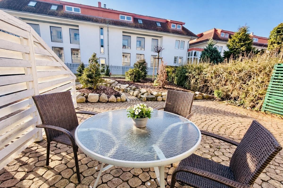 Terrasse mit runden Tisch und drei Stühlen, umgeben von Garten und Wohnhaus.