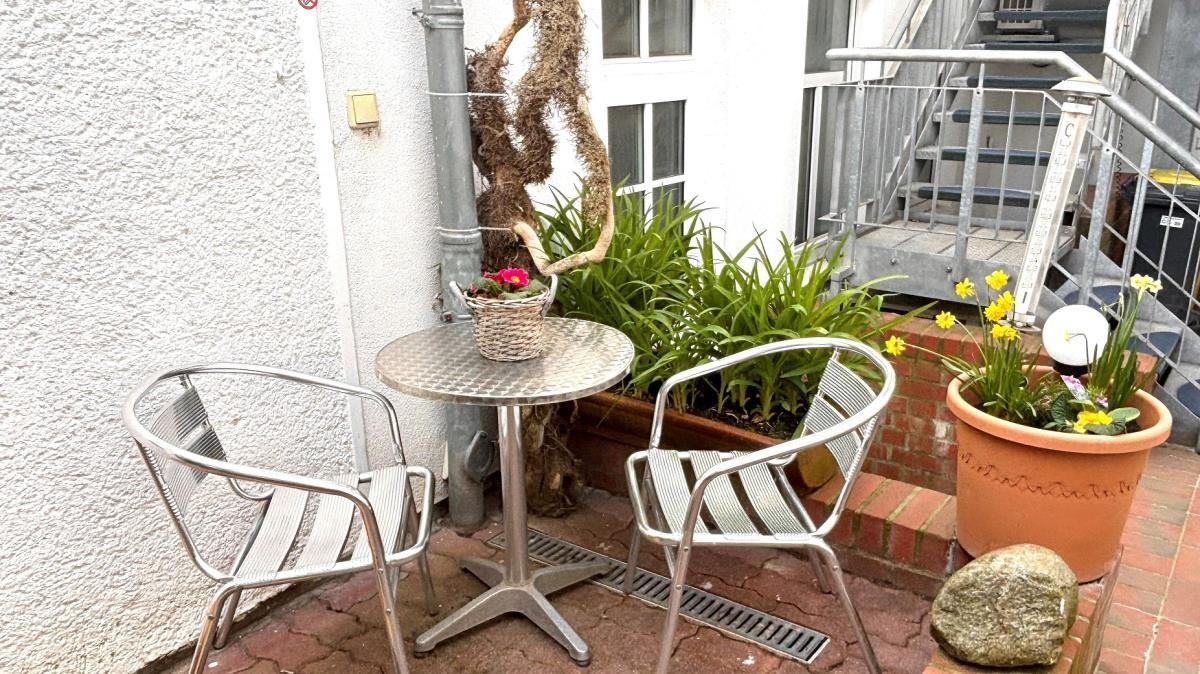 Ein kleiner Balkon mit Tisch, zwei Stühlen und Pflanzen. Hintergrund: Hauswand und Treppe.