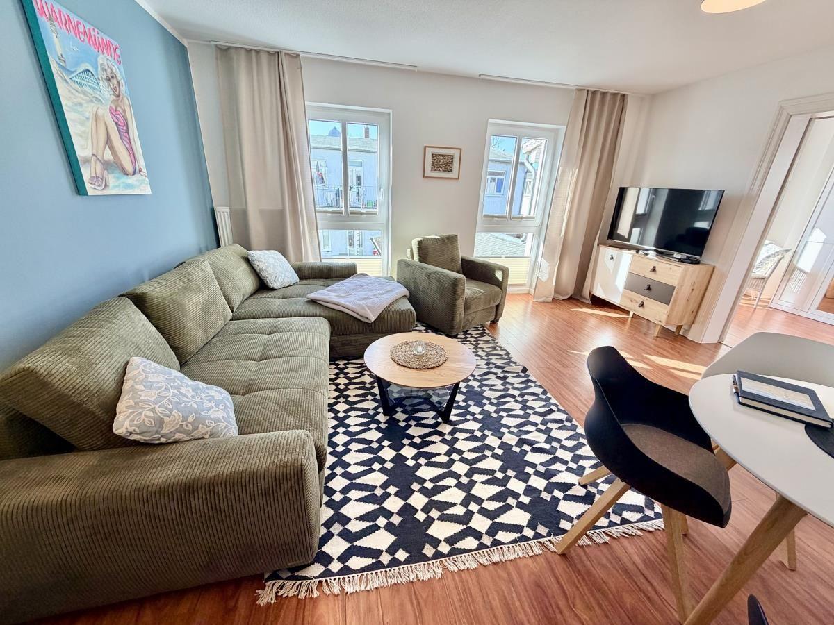 Wohnzimmer mit Sofa, Sessel, TV und Fenster. Teppich und Tisch sind sichtbar.