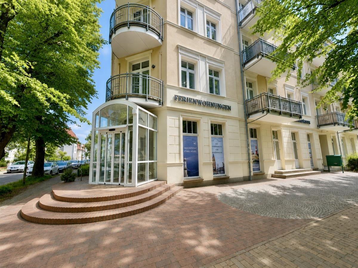 Ferienwohnungen mit Balkonen und Glas-Eingang. Bäume und gepflasterte Fläche vor dem Gebäude.