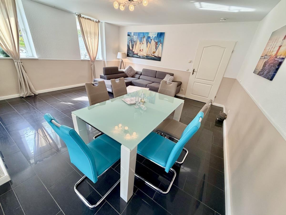 Essbereich mit Glas-tisch und blauen Stühlen. Im Hintergrund Wohnbereich mit Sofa und Wandbild.