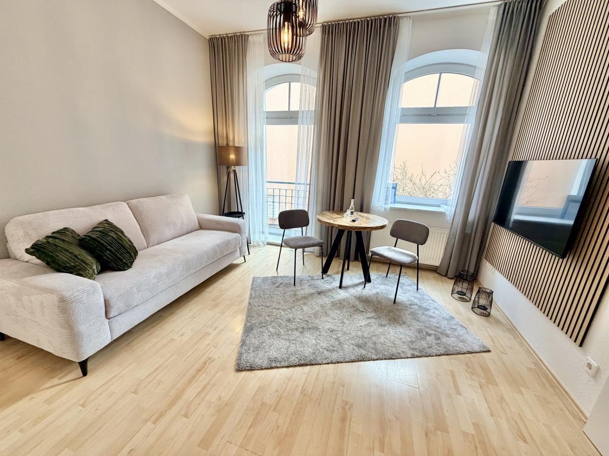 Wohnzimmer mit Sofa, Tisch, Stühlen und TV. Holzboden und große Fenster.