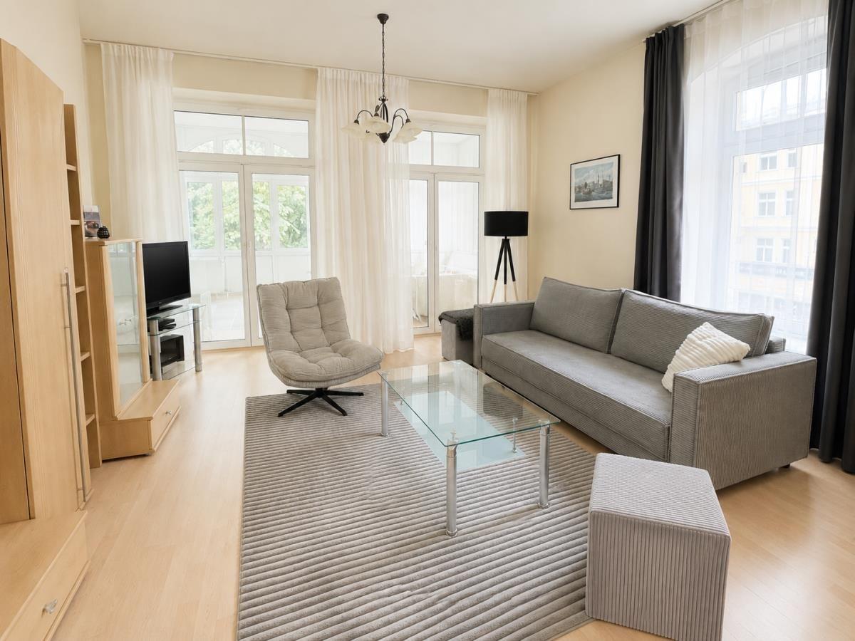 Wohnzimmer mit Sofa, Sessel, Glas-Tisch und großen Fenstern.