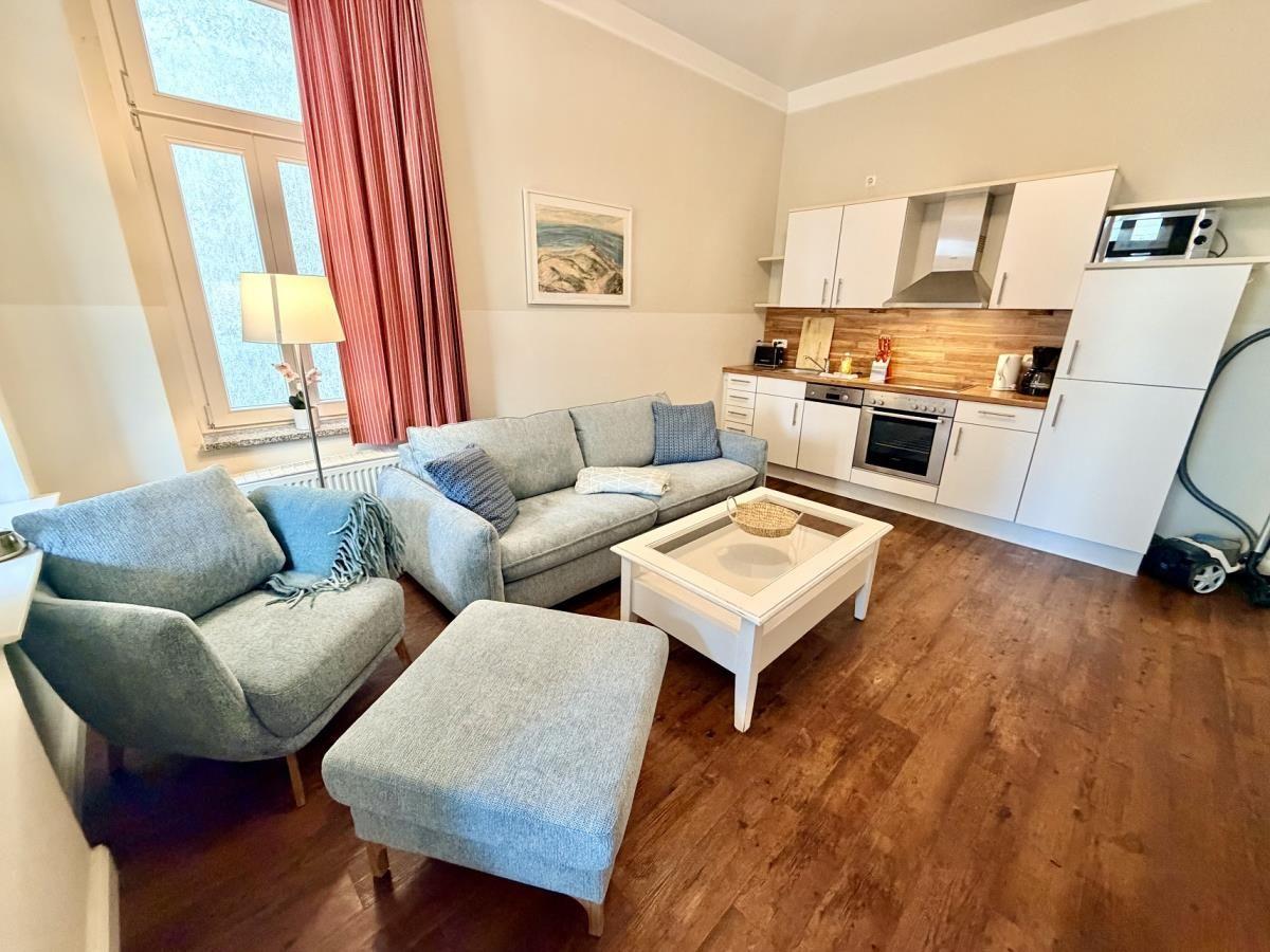 Wohnzimmer mit Sofa, Sessel und Küchenbereich. Holzboden und Fenster mit roten Vorhängen.
