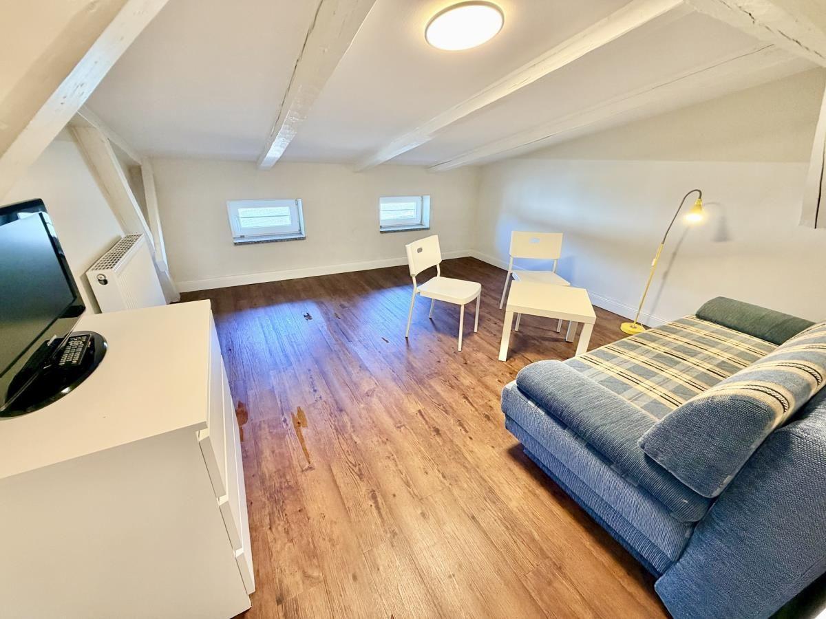 Wohnraum mit Holzboden, Sofa, Tisch, Stühle und TV. Dachboden mit Balken und zwei Fenstern.