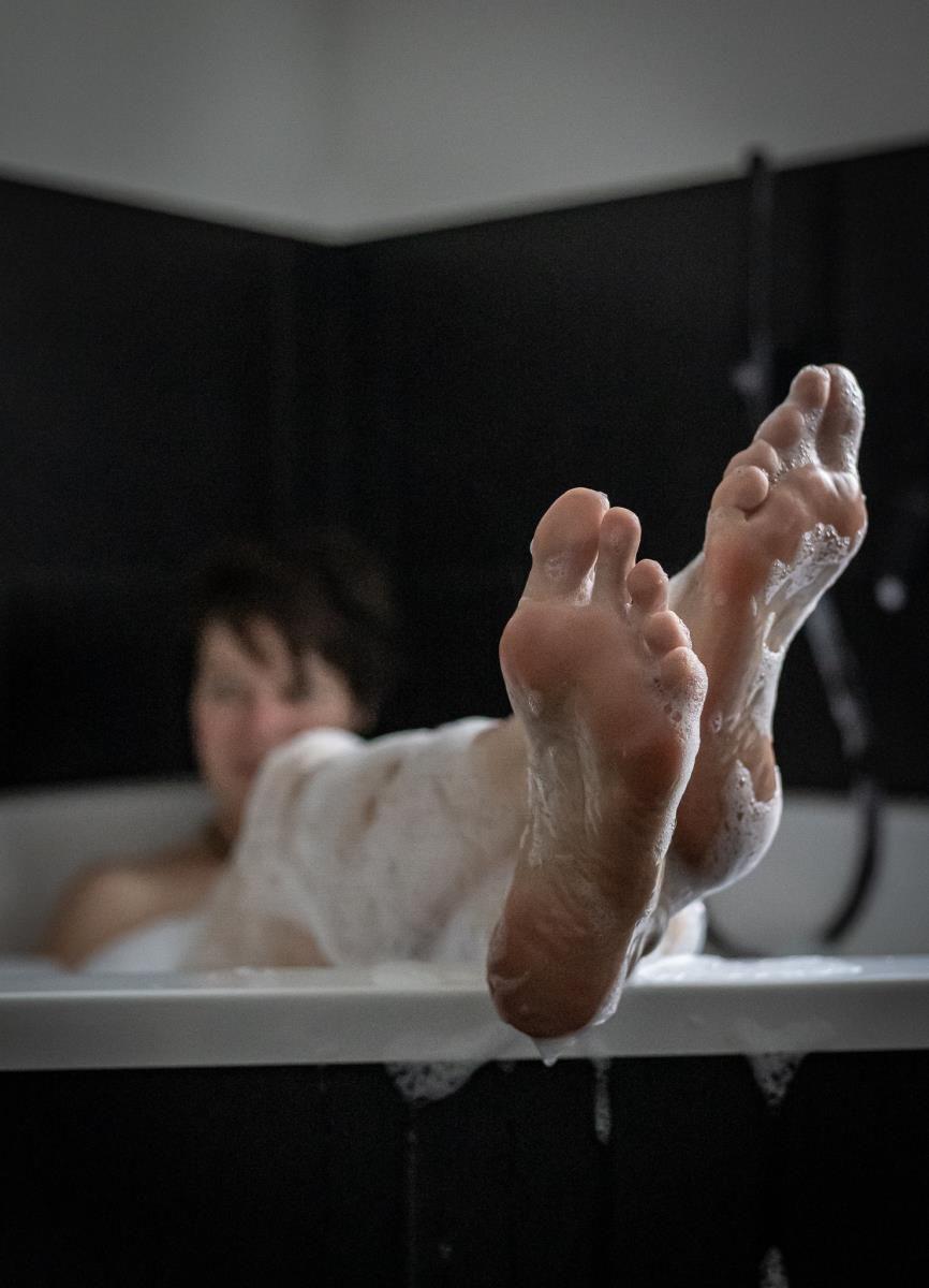 Ein Mensch liegt in einer Badewanne mit Seife und Schaum. Die Füße sind über den Rand hinausgestreckt.