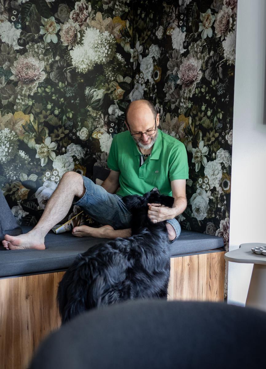 Mann sitzt auf einer Bank vor floraler Wand und streichelt einen schwarzen Hund.