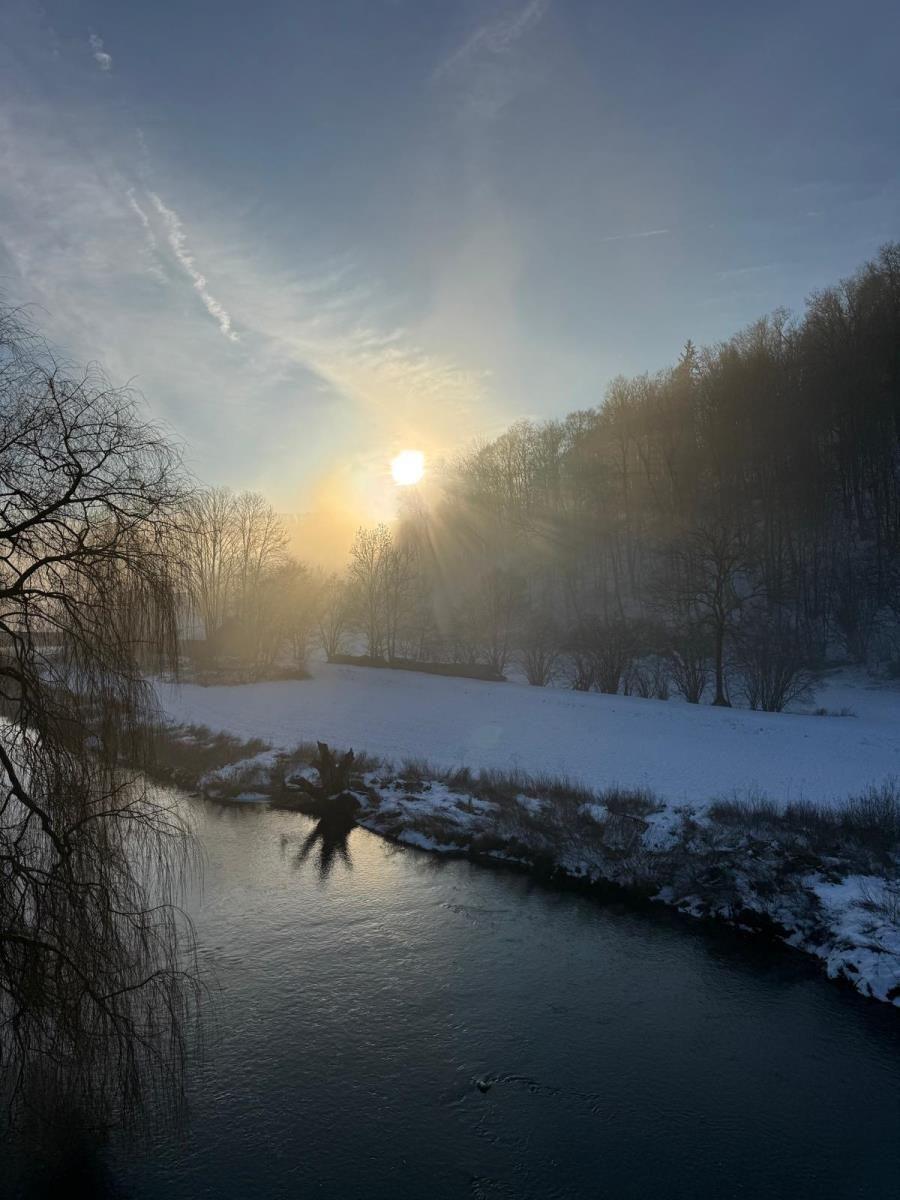 Schneebedeckter Flussuferweg bei Sonnenaufgang mit Bäumen und klarem Himmel.