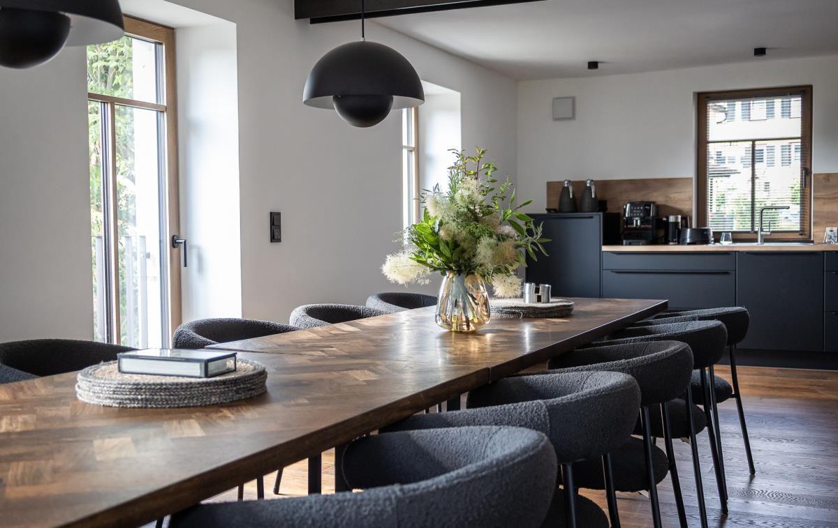 Langes Holztisch mit Stühlen, Blumenvase und Küchenbereich im Hintergrund.