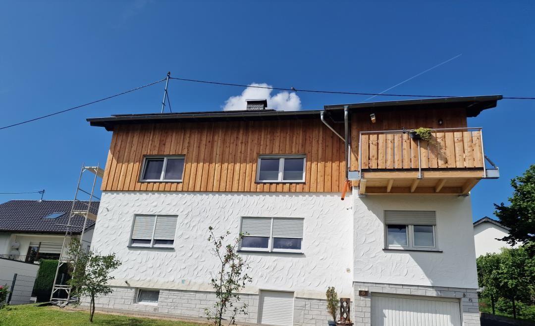 Zweistöckiges Haus mit Holzfassade, Balkon und Garage unter blauem Himmel.