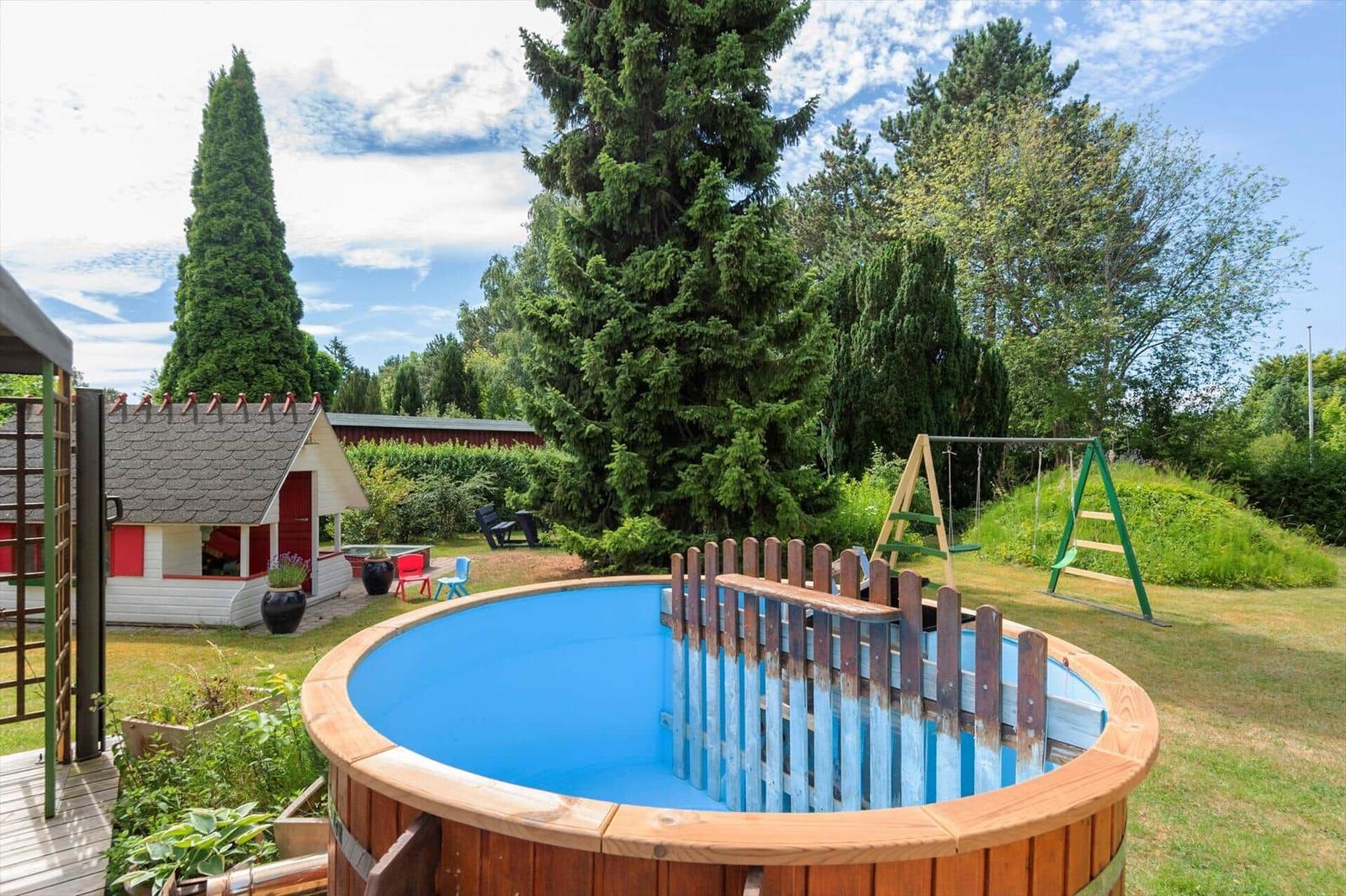 Hochwertiger Holzpool mit Umzäunung. Spielplatz und Gartenhaus im Hintergrund.