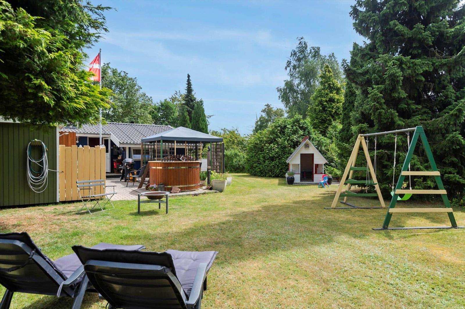 Garten mit Liegen, Whirlpool, Spielhaus und Schaukel. Daneben steht ein Holzhaus mit Flagge.