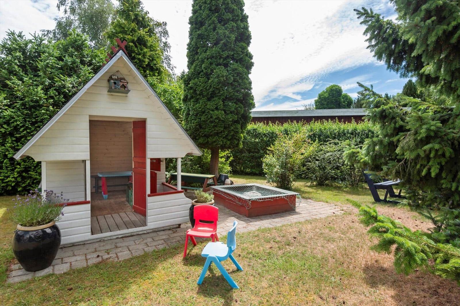 Ein weißer Spielhäuschen mit roter Tür, zwei Kinderstühle und ein Klettergerüst im Garten.
