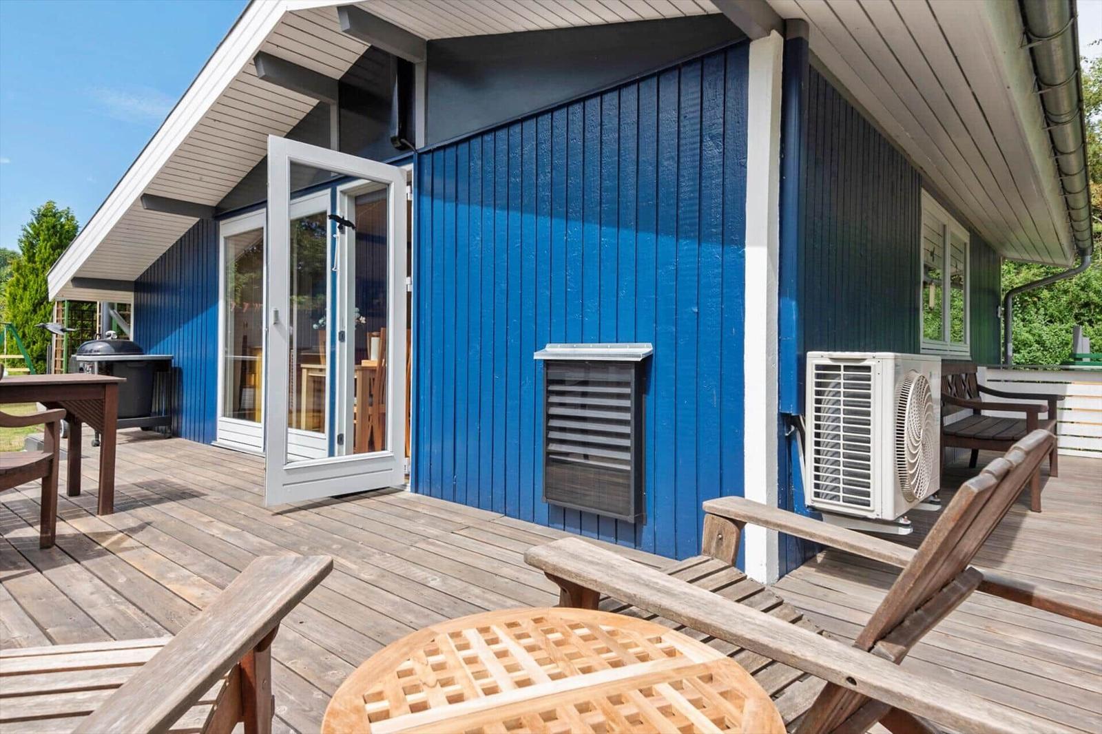 Terrasse mit Holztisch, Stühlen und Grill. Blaue Hauswand mit Schiebetür.
