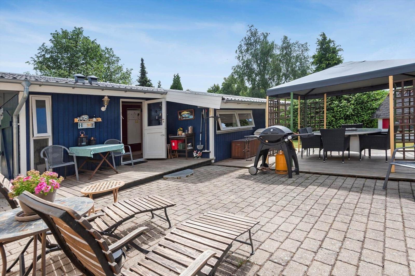 Terrasse mit Grill, Sitzgelegenheiten und blauem Haus. Holzterrasse und Gartenbereich.