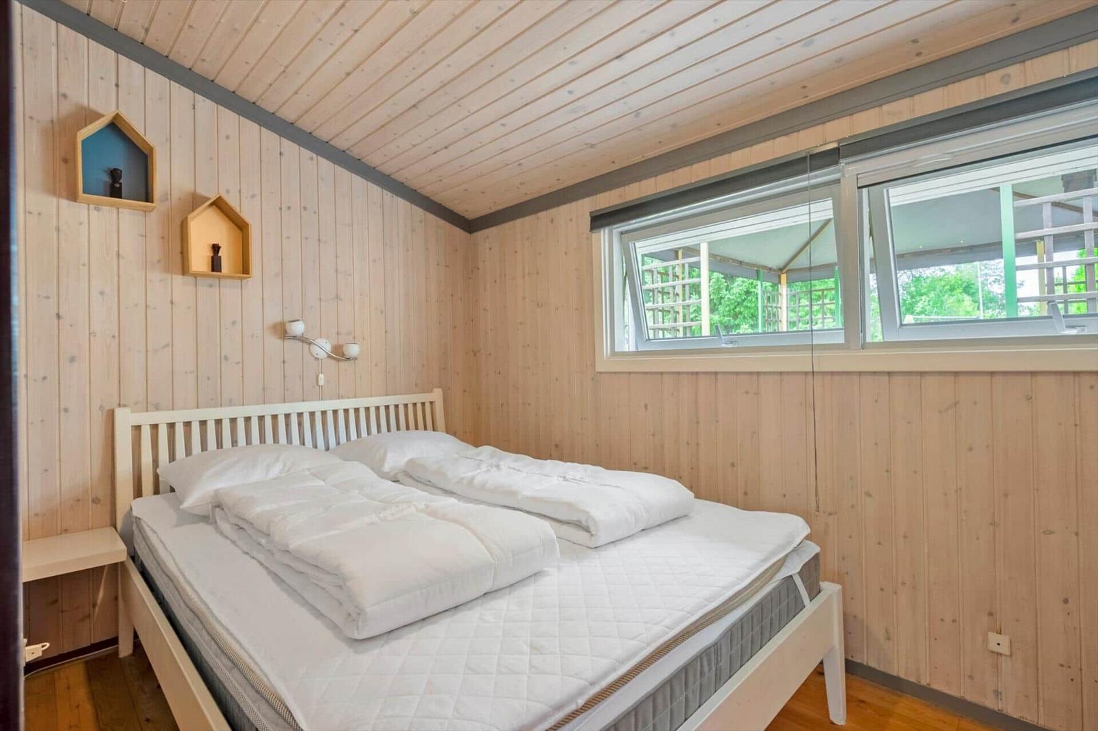 Schlafzimmer mit Bett, Holzwand und Fenster. Zwei kleine Wandregale und eine Nachttischplatte.
