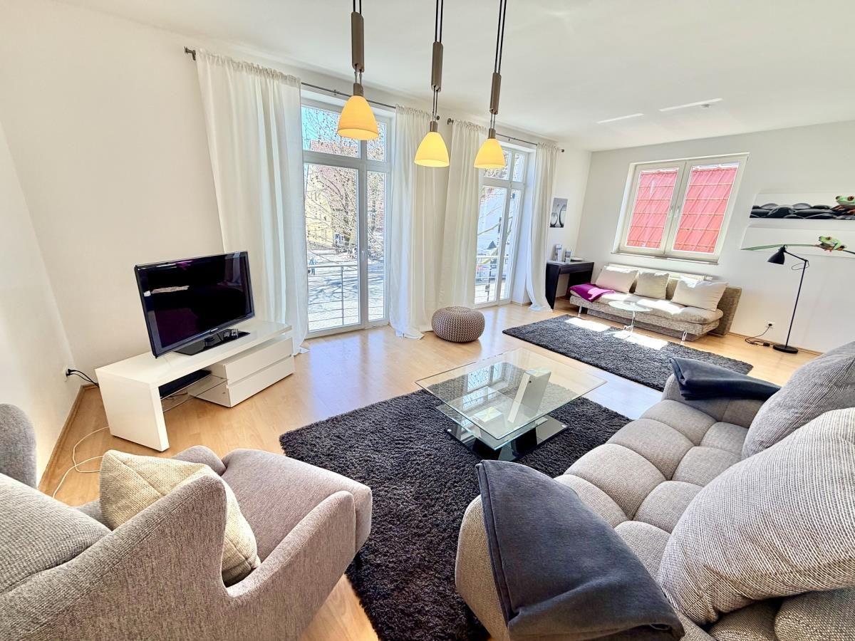 Wohnzimmer mit Sofa, Couchtisch und Fernseher. Große Fenster und helle Wände.