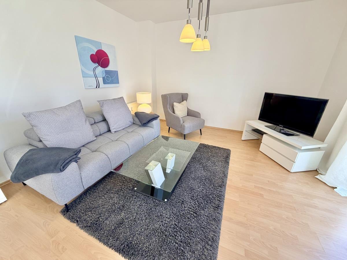 Wohnzimmer mit Sofa, Sessel, Glas-Tisch und Fernseher auf weißem Schrank.