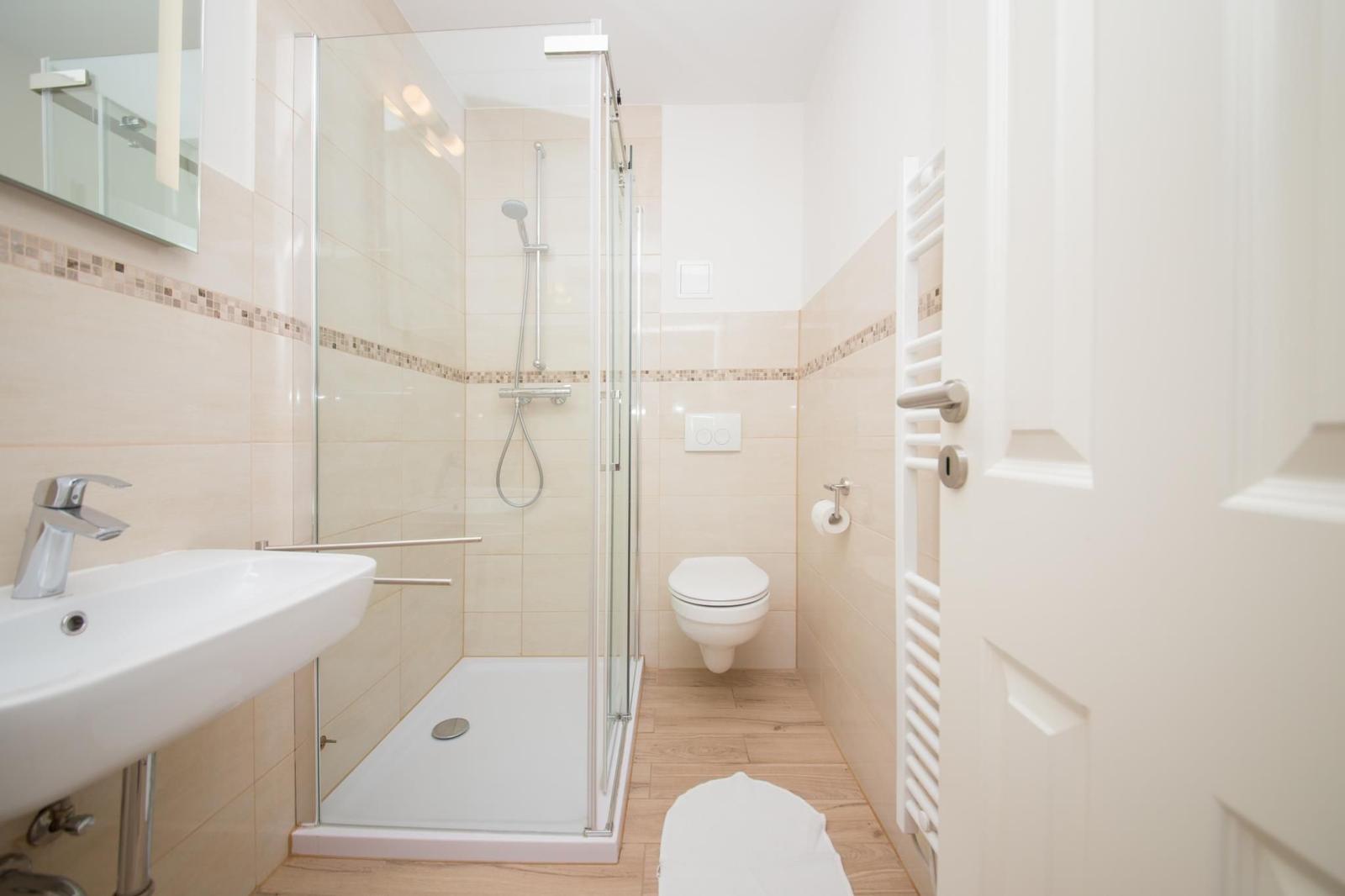 Badezimmer mit Dusche, Waschbecken und Toilette. Holzfußboden und hellbeige Fliesen.