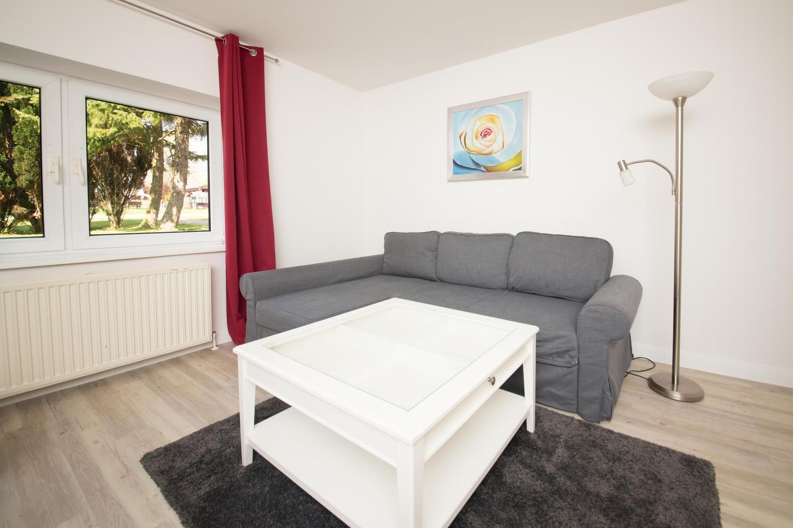 Ein Wohnzimmer mit grauem Sofa, weißem Couchtisch und rotem Vorhang vor dem Fenster.
