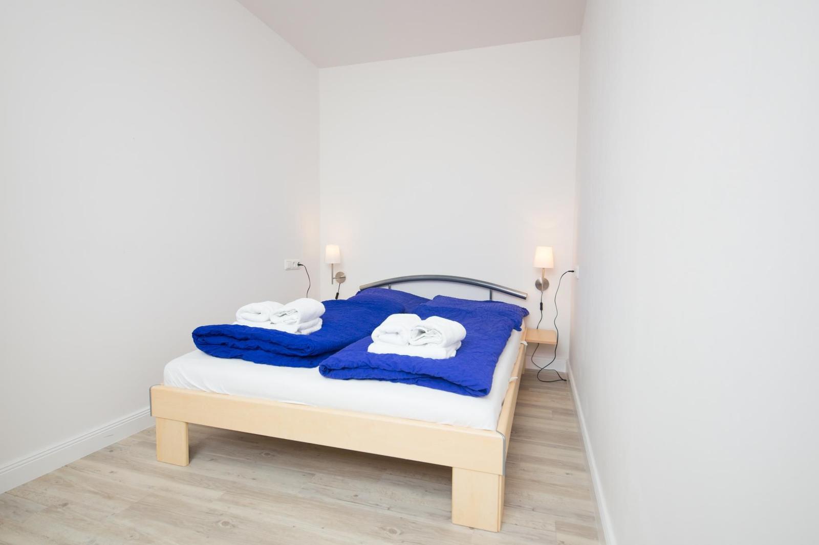 Schlafzimmer mit Bett, blauer Bettwäsche und zwei Wandlampen an den Seiten.