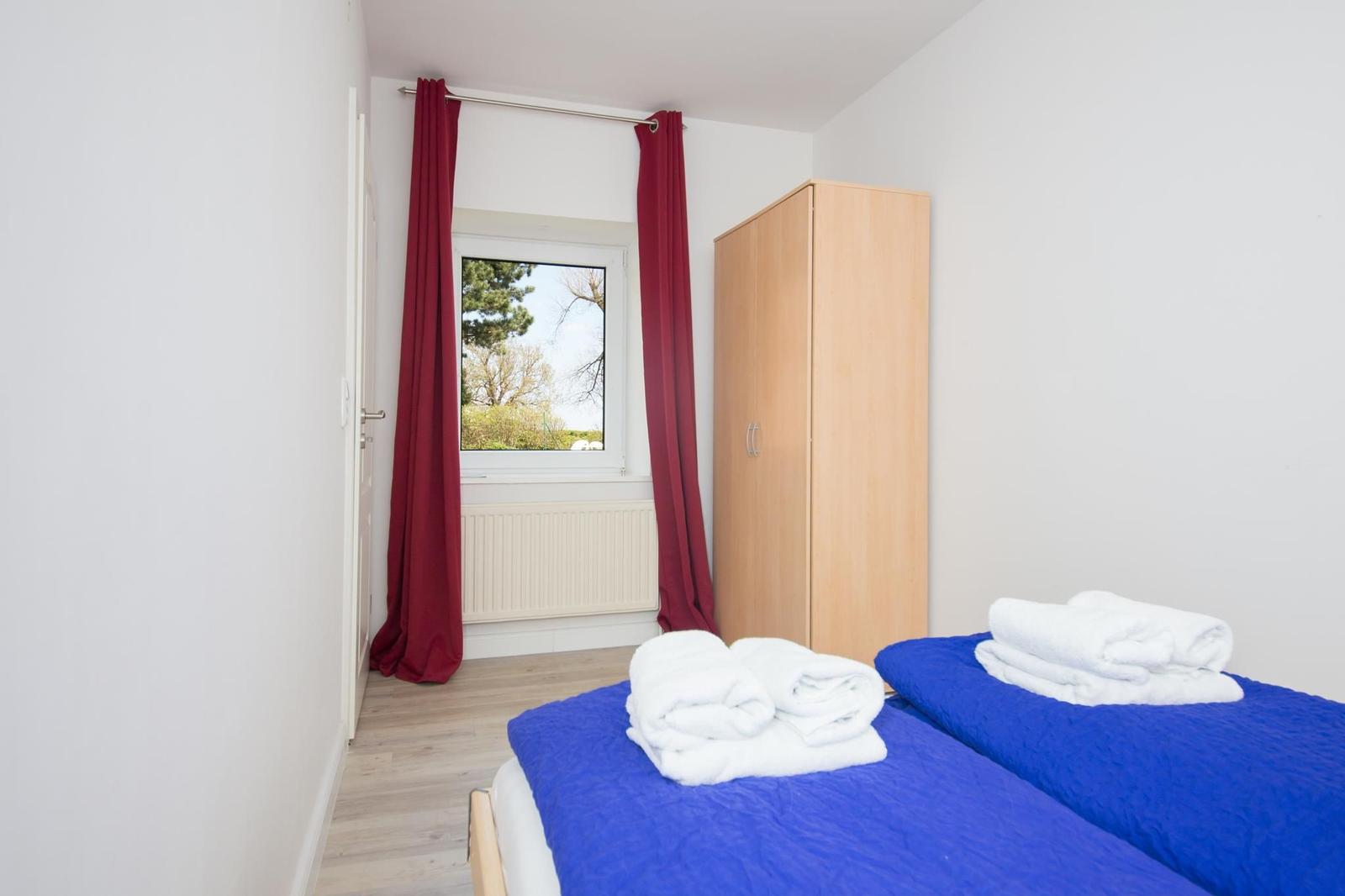 Zimmer mit blauem Bett, roten Vorhängen und Holzschrank. Fensterblick auf Grünflächen.