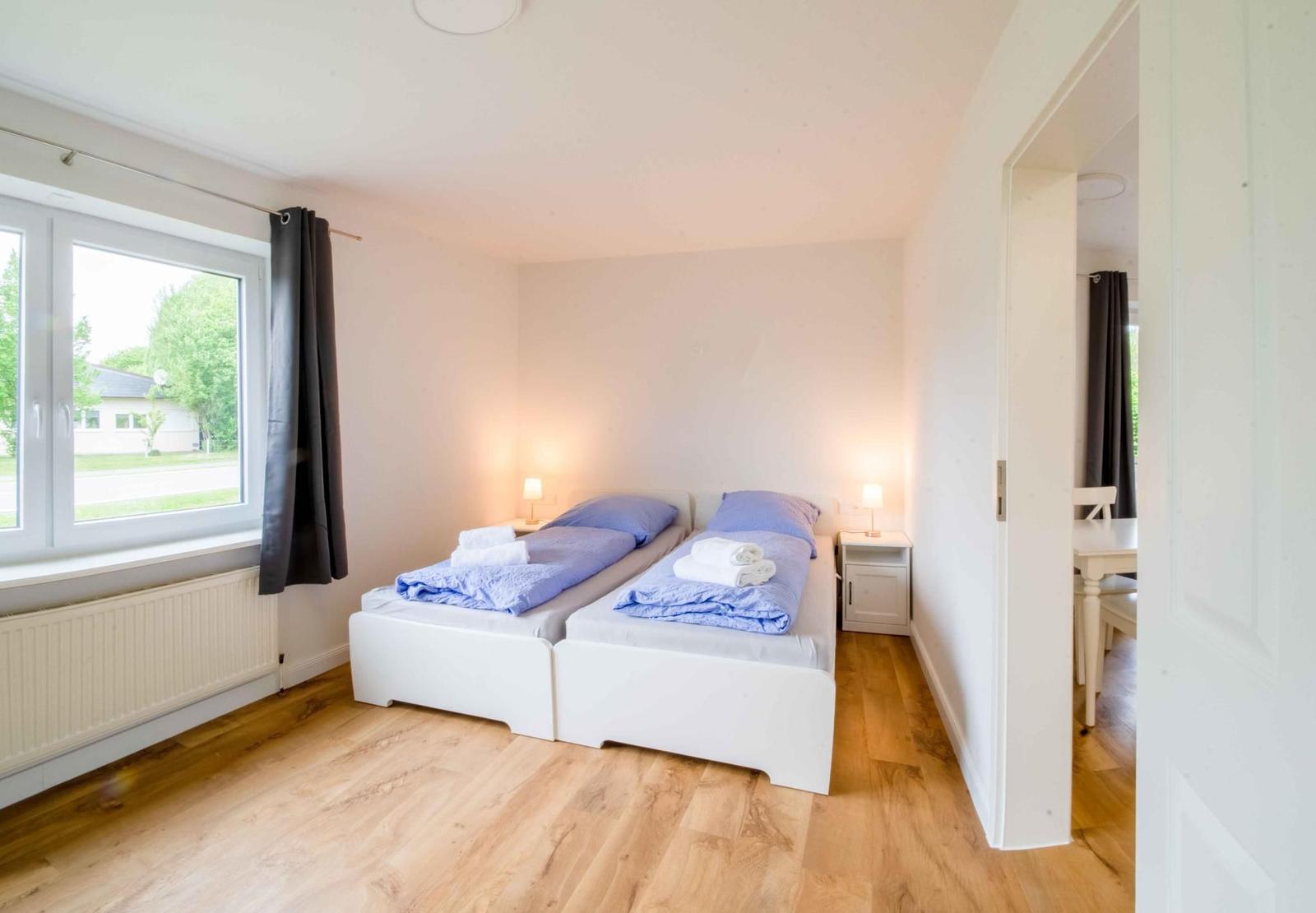 Zwei Betten mit blauen Bettwäschen und Handtüchern. Holzfußboden und Fenster mit Vorhang.