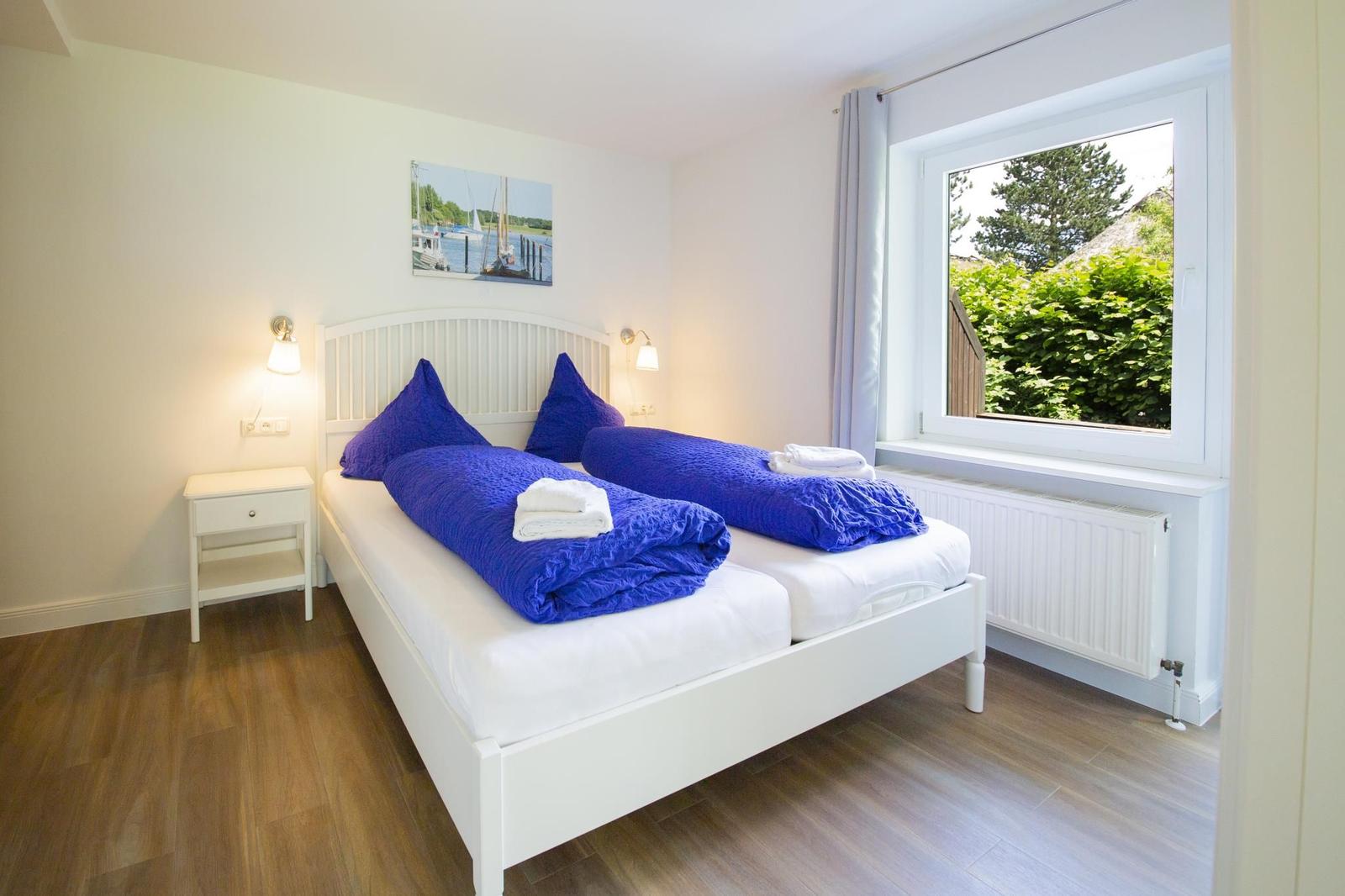 Schlafzimmer mit weißem Bett, blauen Decken und Fensterblick auf Grün