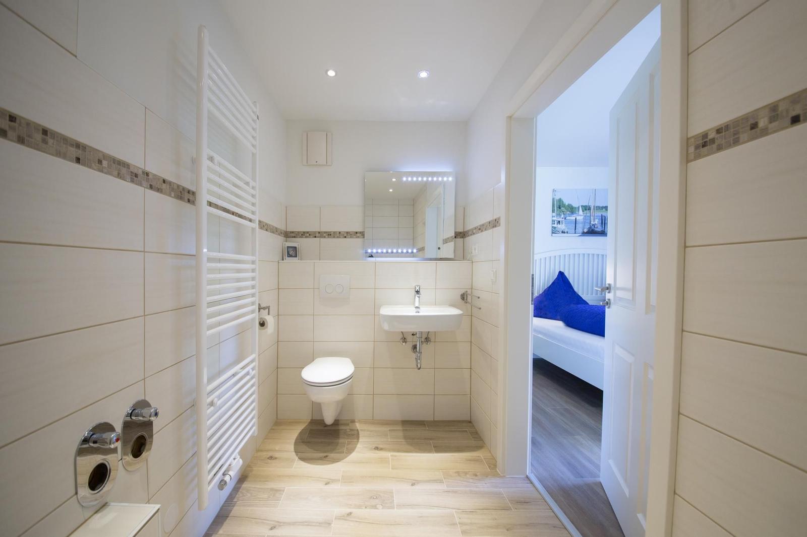 Badezimmer mit Toilette, Waschbecken und Heizkörper. Durchgang zur Schlafzimmer mit blauem Kissen.