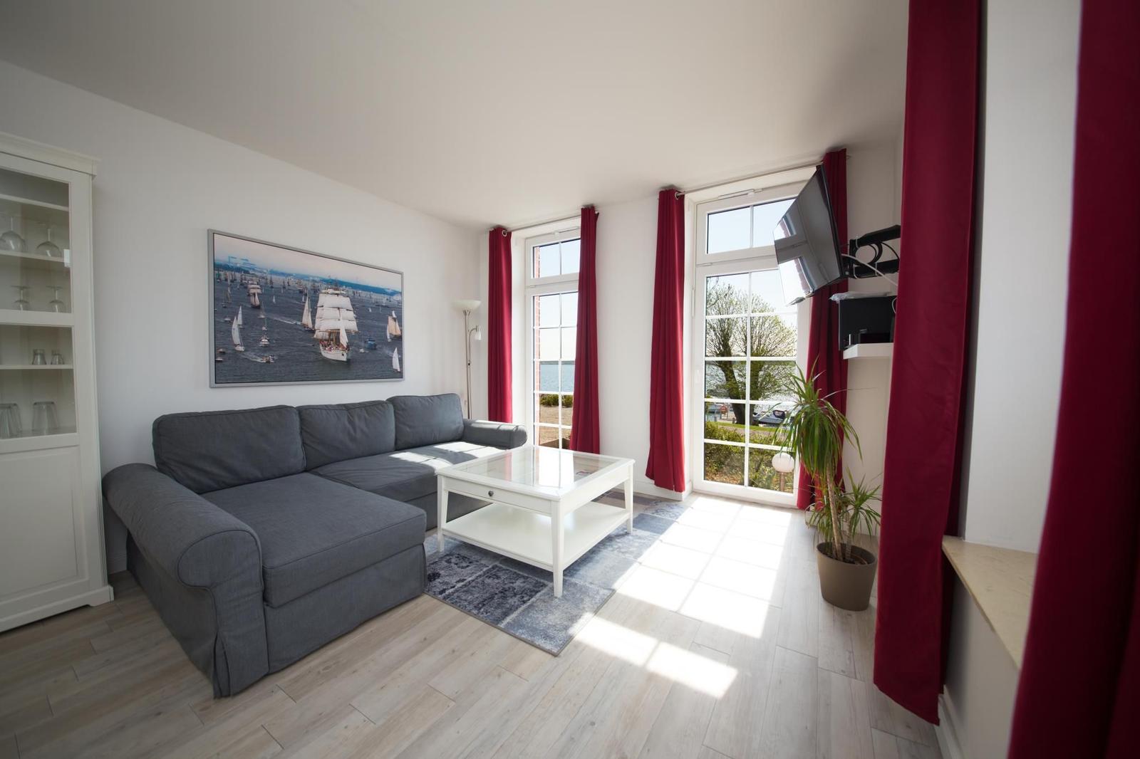 Wohnzimmer mit grauem Sofa, roten Vorhängen und Blick auf den See.