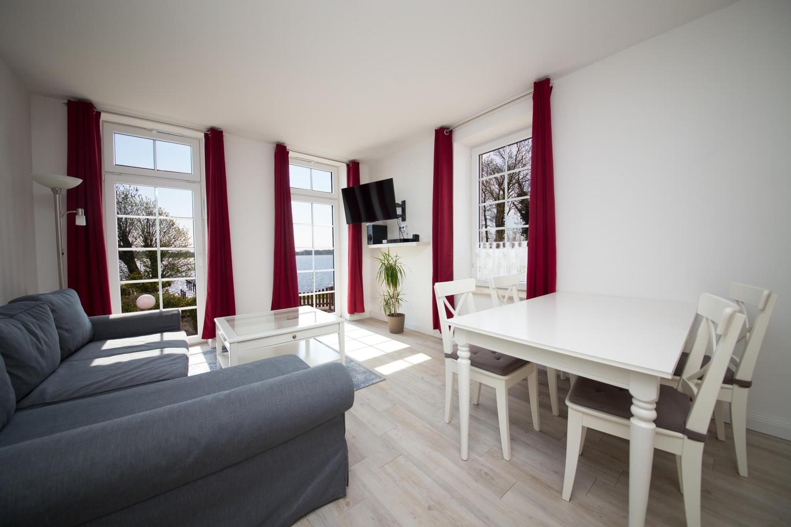 Wohn- und Essbereich mit Sofa, Tisch und Stühlen. Rote Vorhänge und Fenster mit Blick auf Wasser.