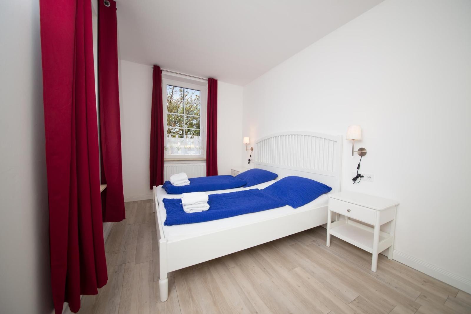 Schlafzimmer mit weißem Bett, blauer Bettwäsche und roten Vorhängen. Holzboden und Nachttisch.