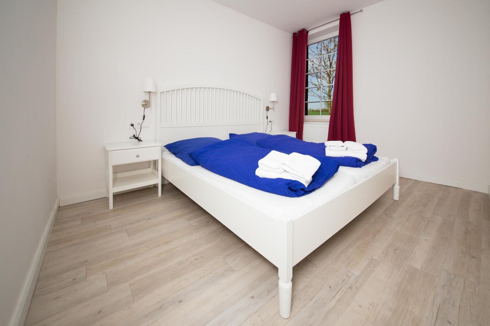 Schlafzimmer mit weißem Bett, blauer Bettwäsche und roten Vorhängen an einem Fenster.