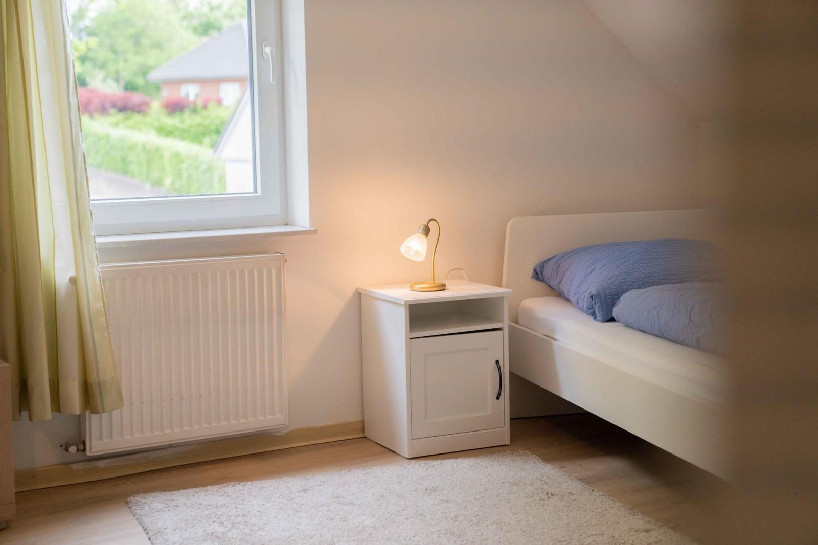 Ein Schlafzimmer mit Bett, Nachttisch und Fenster. Auf dem Nachttisch steht eine Lampe.