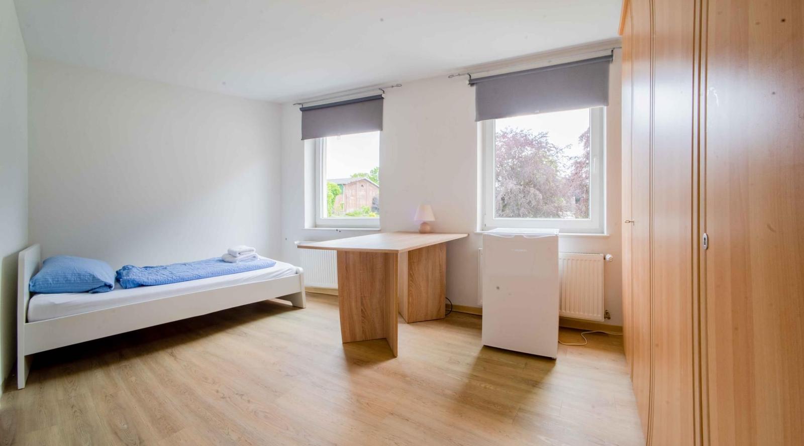 Schlafzimmer mit Bett, Schreibtisch, Kleiderschrank und zwei Fenstern.