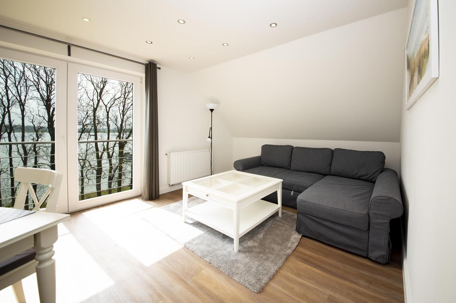 Wohnzimmer mit grauem Sofa, weißem Tisch und Balkontür zum See.