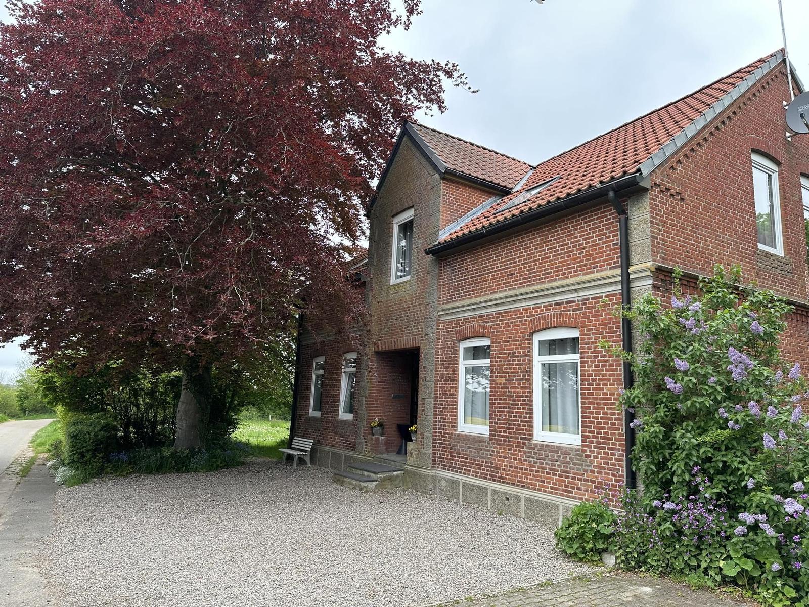 Rotes Ziegelhaus mit Garten, Baum und Blumenhecke. Eingangstreppe und Bank vor dem Haus.